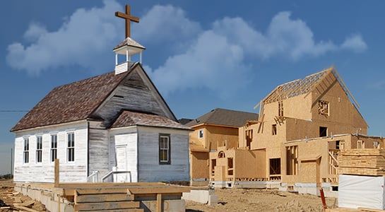 An old white wooden church with a cross stands adjacent to a partially constructed house under a clear blue sky.