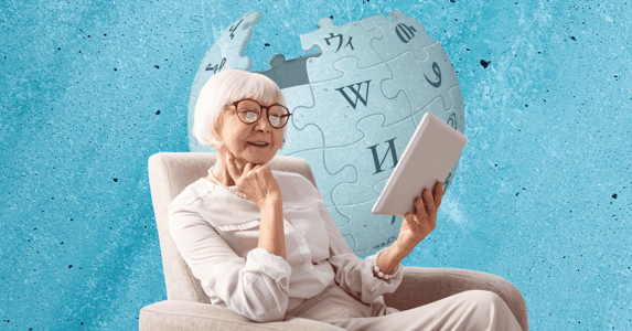 A woman with white hair and glasses sits in a chair while reading a tablet.