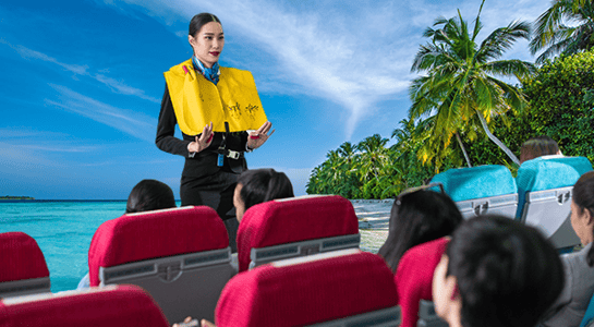Indoor scene: audience in red chairs, person in yellow life vest addressing them; backdrop of tropical coastal scene, indicating a travel or tourism-related safety briefing.