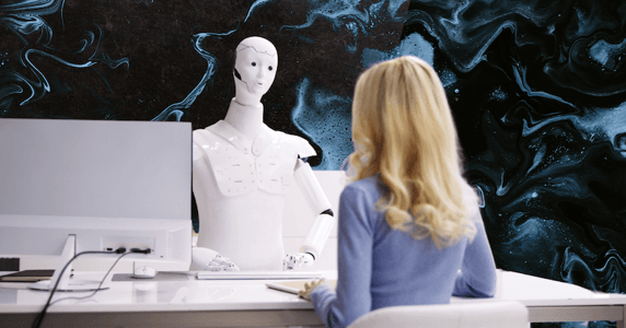 A blonde woman in a blue sweater sits at a desk opposite a white humanoid robot.