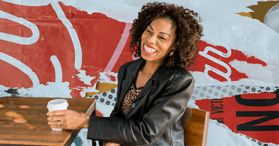 A woman smiling and holding a coffee sitting at a table.