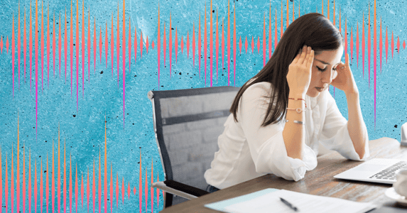 A woman with black hair and a white shirt leans on a desk with her eyes closed and her head in her hands.