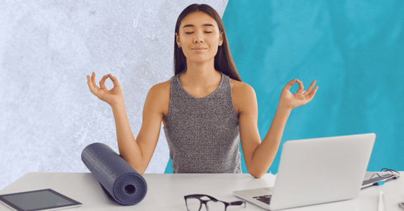 A woman meditating at her desk with a yoga mat on one side and a laptop on the other. 