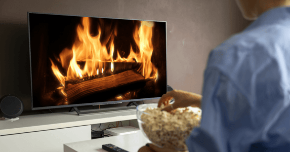 A person eating a bowl of popcorn and looking at a TV that’s playing a fireplace. 