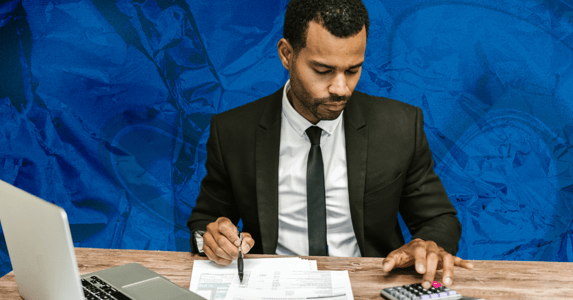 A young Black accountant sits at a desk with a laptop and several papers. He is wearing a suit, holding a pen, and using a calculator.