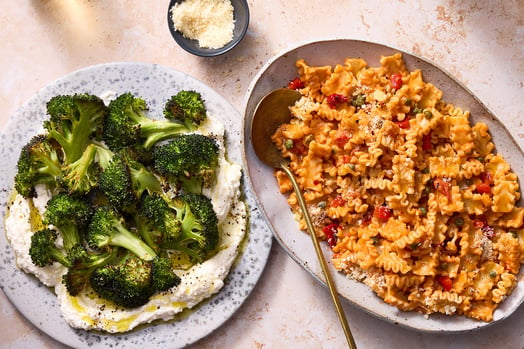 A plate of broccoli next to a plate of pasta and a small cup of parmesan cheese. 