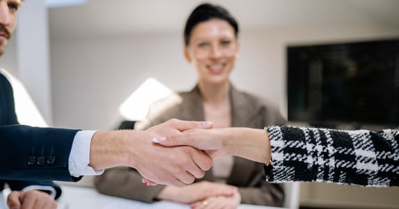 A man and a woman shake hands. In the background, a woman is smiling. 