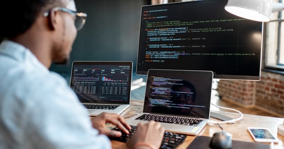 A Black man sits behind a laptop and two screens, writing code.