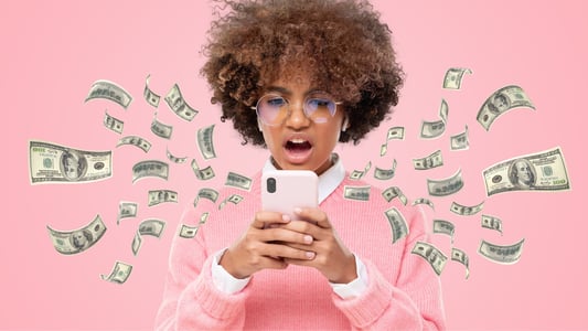 A young Black woman in a pink sweater and glasses looks at her phone in disbelief as money flies out.