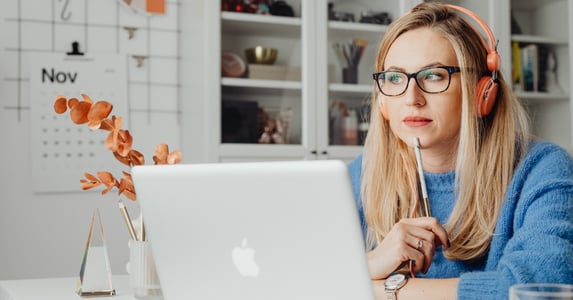 A white woman sits alone at a desk in front of her laptop. She wears a blue sweater, glasses, and headphones.