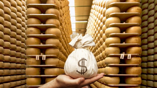 A hand holds a sack of money in front of a large collection of parmesan cheese wheels