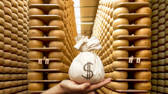 A hand holds a sack of money in front of a large collection of parmesan cheese wheels
