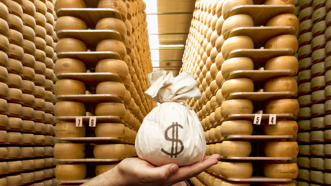 A hand holds a sack of money in front of a large collection of parmesan cheese wheels