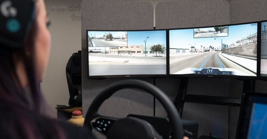 A woman sits behind two monitors depicting a roadway and a steering wheel.