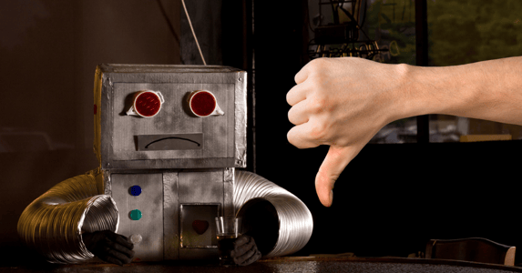 A robot with a frown sits at a table in front of a person's hand making a thumbs down sign.
