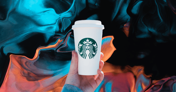 A hand holds up a Starbucks cup against a blue and peach background.