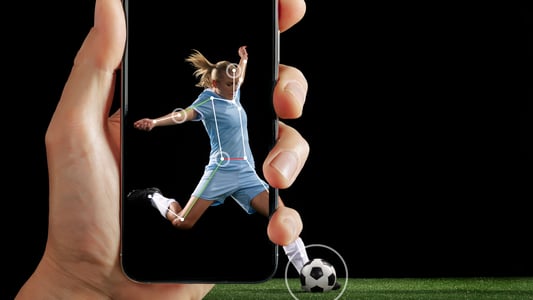 A hand holds up a phone depicting a woman kicking a soccer ball as an AI system tracks her movements. 