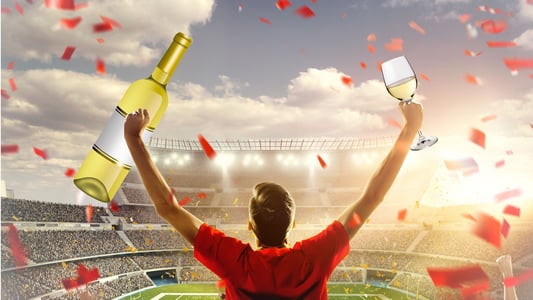 A fan at a stadium holding a bottle and glass of wine in the air. 