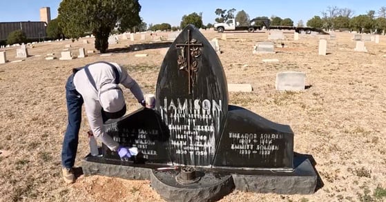 A man in a gray hoodie and denim overalls scrubs a headstone with a sponge.