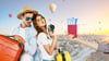 A vacationing couple holds a suitcase and camera. In the distance, several hot air balloons rise over a city. In the foreground, a hand offers two tickets.
