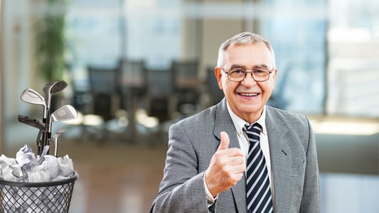 A senior man in a suit, tie, and glasses gives a thumbs up while standing next to a trash bin containing a set of golf clubs.