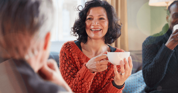 A smiling woman in a sweater holding a cup of tea sits chats with two men.