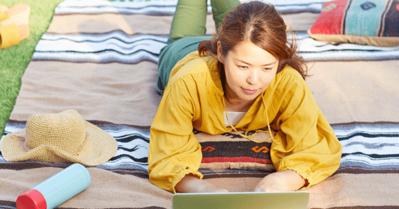 A young Asian woman lies on her stomach on a blanket in a park while working on her laptop.